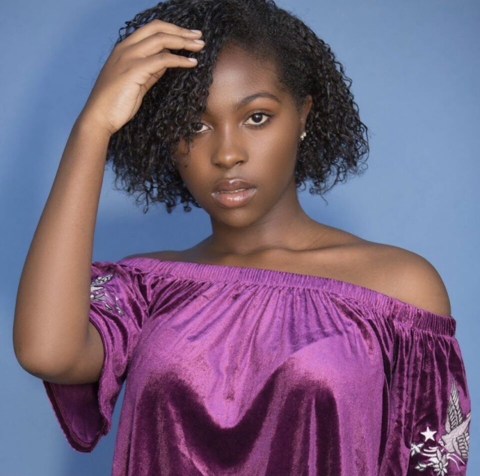 Young woman with natural curly hair wearing an off-shoulder purple top.