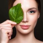 Woman holding a green leaf over one eye, symbolizing natural beauty.