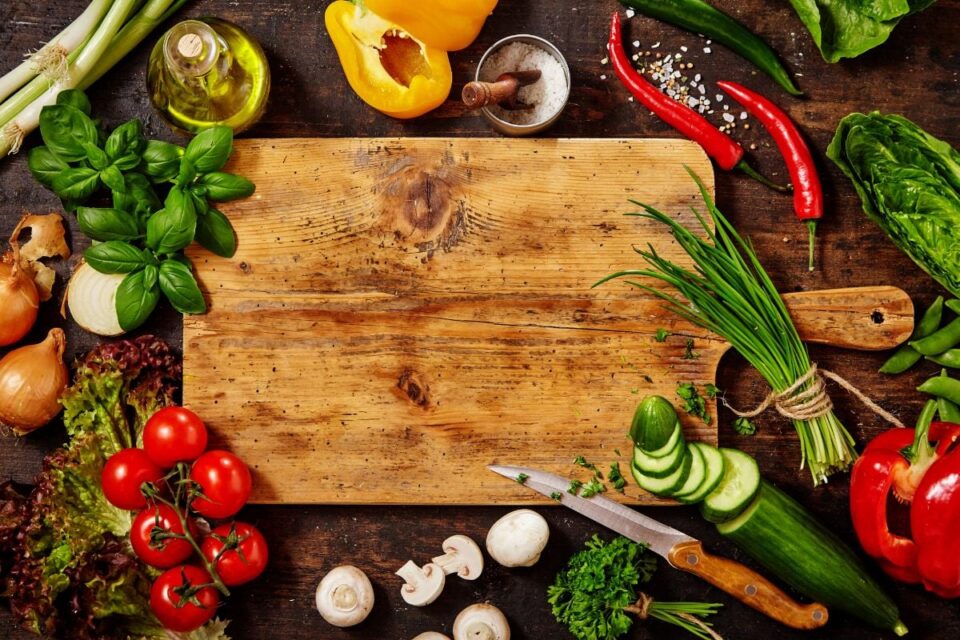 Wooden cutting board surrounded by fresh vegetables and herbs.