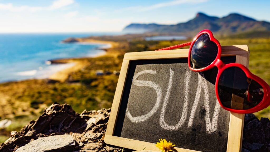 Sunglasses resting on a chalkboard with 'SUN' written, overlooking a sunny coastal landscape.