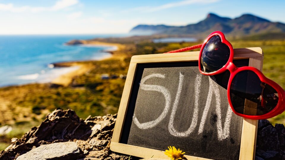 Sunglasses resting on a chalkboard with 'SUN' written, overlooking a sunny coastal landscape.
