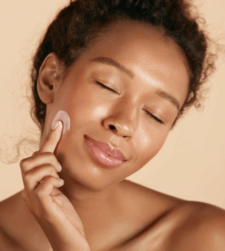 Woman gently applying cream to her cheek with eyes closed.